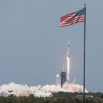 A SpaceX Falcon 9 rocket lifts off, framed by an American flag atop a flagpole.