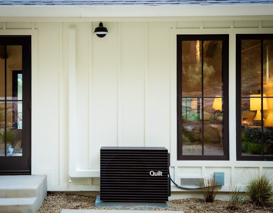 A Quilt heat pump outdoor unit sits outside a white house.