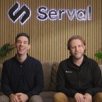 Two caucasian men sit on a couch with a company logo behind them.