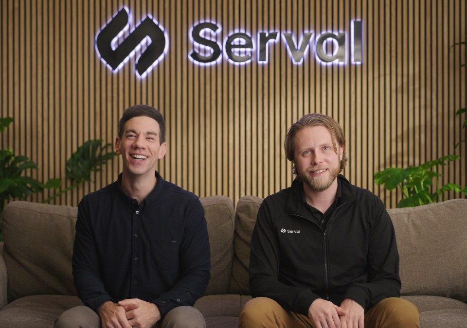 Two caucasian men sit on a couch with a company logo behind them.