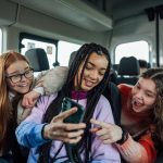 A front view of a group of three teenage girls on the minibus on the way to go on a hiking field trip. They are looking at one of the girls mobile phones and smiling and pointing at the screen.