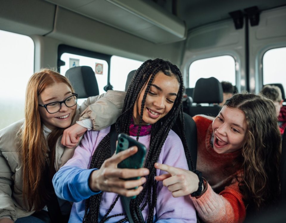 A front view of a group of three teenage girls on the minibus on the way to go on a hiking field trip. They are looking at one of the girls mobile phones and smiling and pointing at the screen.