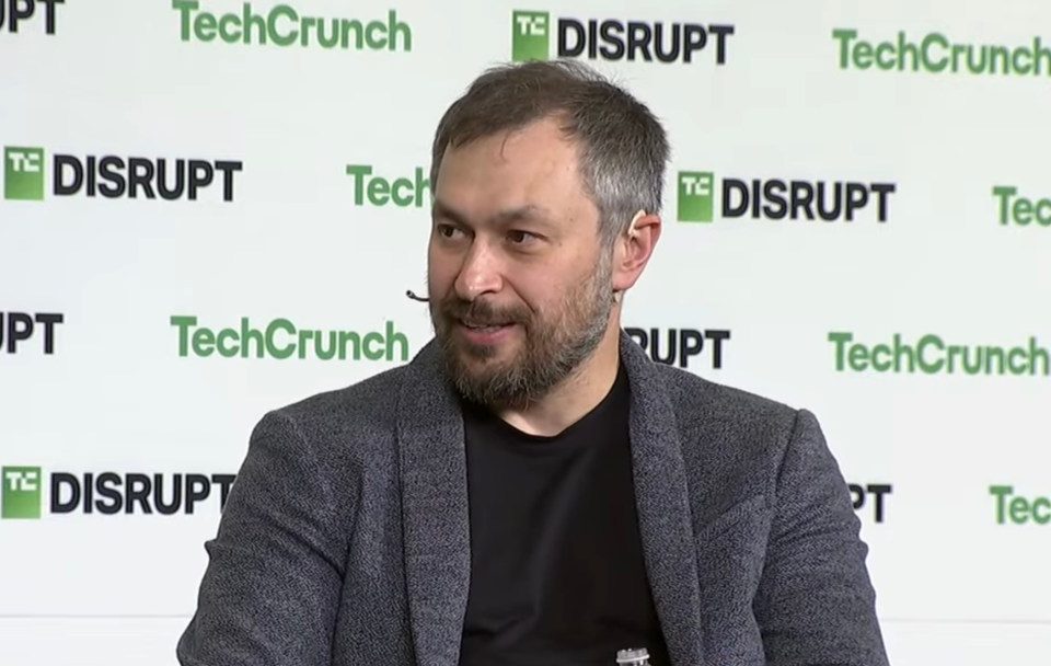 A bearded man grins against a TechCrunch-branded backdrop